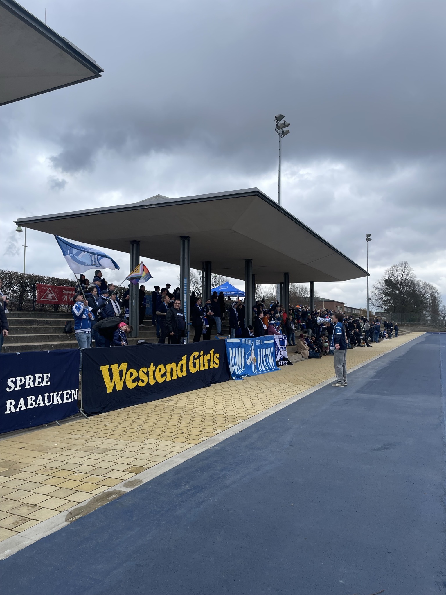 Blick auf supportende Fans. Im Vordergrund hängen die Banner der Fangruppen der Hertha Frauen (Donna Hertha, Westend Girls, Spreerabauken)