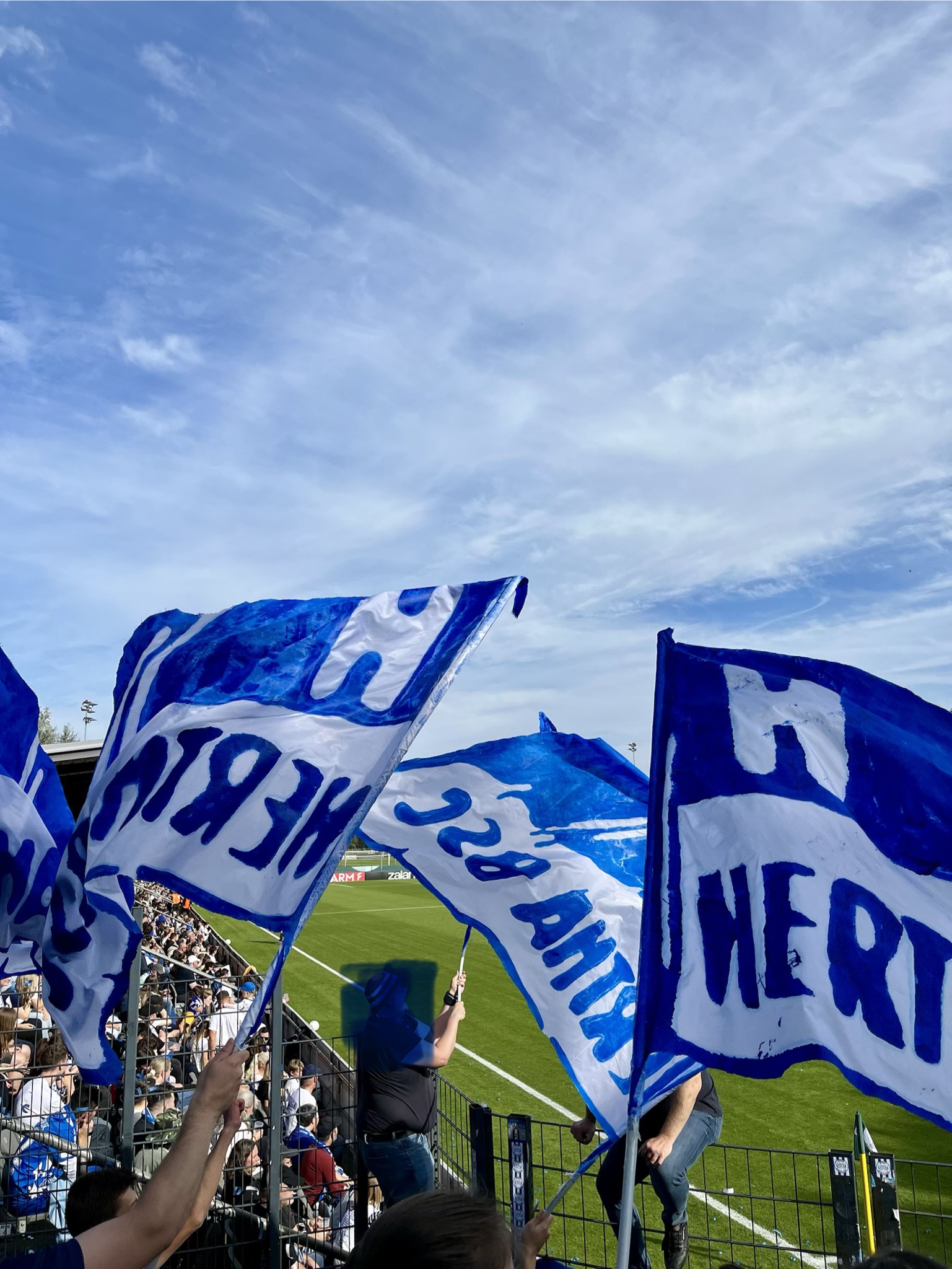 Blau-weiße Fahnen wehen im Wind. Im Hintergrund das Spielfeld